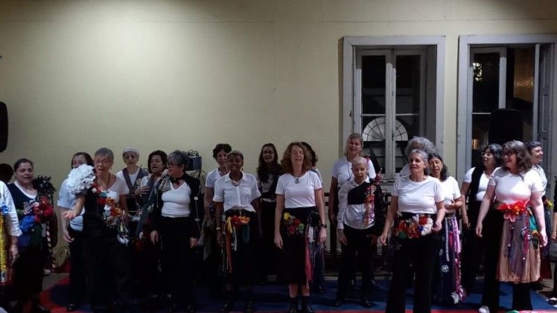 Mulheres de diferentes etnias, com camisetas brancas e aplicações de tecidos coloridos, cantando similar a um coral