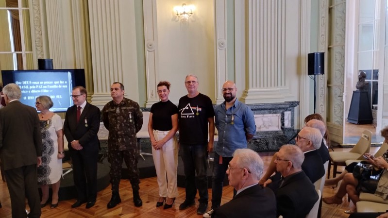 Sete pessoas estão em pé diante de um público sentado, numa sala do Palácio Piratini com arquitetura de estilo clássico. As pessoas que estão de pé, ostentam uma medalha colocada ao peito. Entre elas esta nossa diretora Doris Couto vestindo blusa preta e calça branca, com os cabelos ruivos arrumados num coque, ao seu lado Claus Farina, está de blusa e calça preta.  
