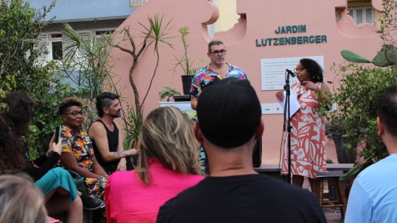 A imagem mostra uma apresentação do Sarau Livre no Jardim Lutzenberger, na Casa de Cultura Mario Quintana. No palco estão em pé uma mulher (ao microfone) e um homem, ladeados por uma mulher e um homem sentados. O público, sentado, assiste ao sarau