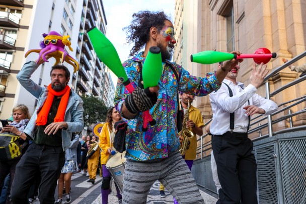 Imagem de pessoas em cortejo na Rua Duque de Caxias, com malabares e instrumentos musicais. 