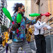 Imagem de pessoas em cortejo na Rua Duque de Caxias, com malabares e instrumentos musicais. 
