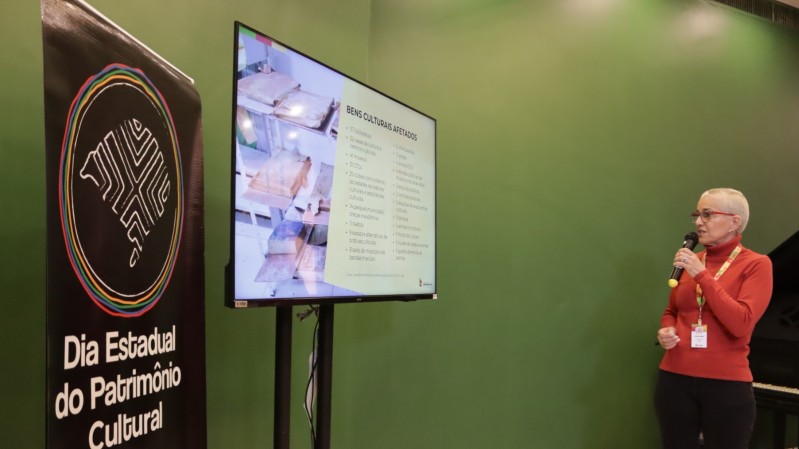 Secretária Beatriz Araujo diante de um monitor com relação de instituições culturais afetadas pelas enchentes no Rio Grande do Sul. Ao fundo, parede em tom de verde e ao lado esquerdo, banner com logo do Dia do Patrimônio Cultural. 