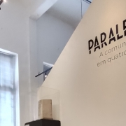 Parede branca divulgando a exposição ''Paralelos: A comunicação em quatro linhas''.