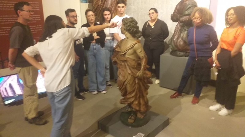 No centro da imagem escultura de madeira representa um anjo esculpido em madeira que é observado por um grupo de adultos enquanto mediadora aponta um detalhe.