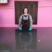 Leonardo Tavares, homem branco com cabelo castanho escuro na altura dos ombros, usando luvas de borracha. Ele está ao centro da imagem, em frente a uma janela fechada, com água da enchente até a altura do quadril. Na água, é possível notar folhas e outras impurezas boiando.
