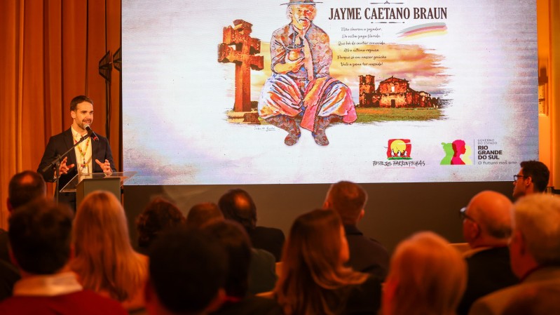 Leite falando ao microfone no lançamento dos Festejos Farroupilhas 2024. Parte da plateia pode ser vista de costas e há uma projeção em uma tela com a imagem de Jayme Caetano Braun, homenageado deste ano.