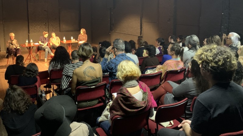 Teatro com diversas pessoas na plateia assistindo a um seminário, com quatro pessoas no palco sentados em cadeiras e duas mesas ao centro dividindo as palestrantes.