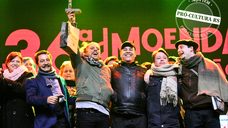 No palco da Moenda da Canção, vários homens estão em pé, sorrindo e de frente para o público. Um deles ergue um troféu com uma das mãos. Sobre a imagem há um selo, no qual está escrito “Aqui tem Pró-Cultura RS”.