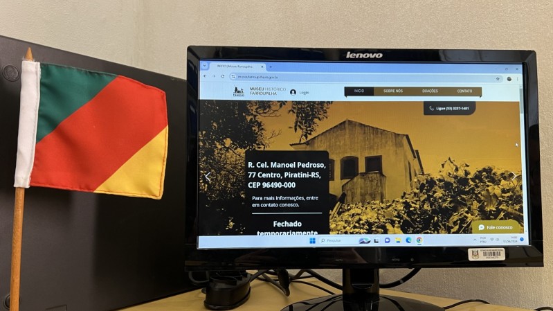 Ao lado esquerdo, uma bandeira em miniatura do Estado do Rio Grande do Sul, nas cores verde, vermelho e amarelo, respectivamente. Ao lado direito, um monitor de computador, com o novo site do Museu Histórico Farroupilha, com um fundo amarelado, um monumento histórico do Estado ao centro, com as informações sobre o Museu.