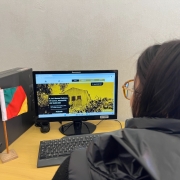 Ao lado esquerdo, uma miniatura da bandeira do Rio Grande do Sul, ao centro, um computador com a tela inicial do site do Museu Histórico Farroupilha, com um fundo amarelado e com um monumento do Estado. Ao lado direito, uma mulher com cabelos castanhos e óculos observando o monitor do computador.