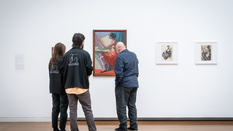 Um homem observa um quadro em uma exposição. Ao seu lado, há um homem e uma mulher, ambos com um uniforme no qual está escrita a palavra "Educativo"