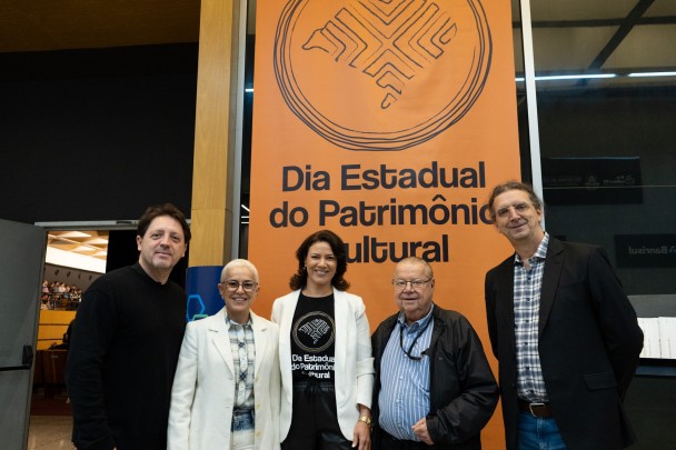 Cinco pessoas em pé, diante do banner do Dia do Patrimônio