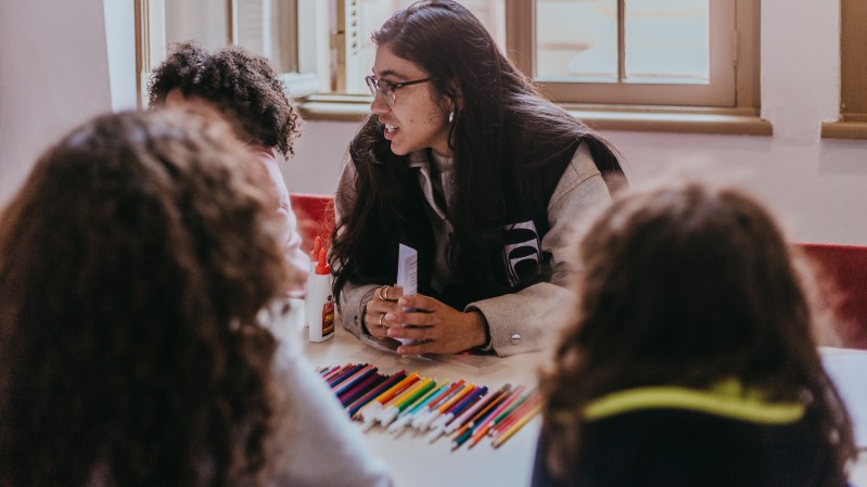 Uma mulher ministra uma oficina para crianças, todos sentados em uma mesa, na qual há materiais escolares