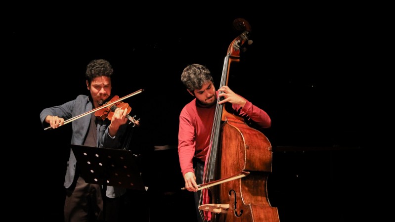 Um músico toca violino e, ao seu lado, outro músico toca violoncelo