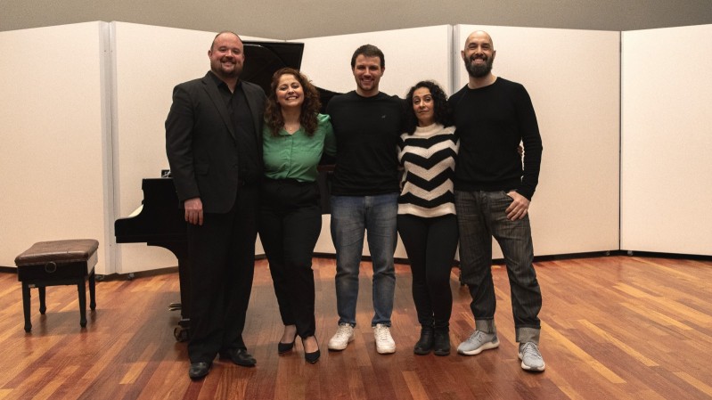 Imagem do Grupo "QuattrUm". Ao fundo, um teatro com paredes brancas. Na imagem, 5 pessoas, um homem branco, de cabelos raspados, usando roupas pretas. Em seu lado direito, uma mulher branca, de cabelos cacheados, usando camisa social verde e uma calça preta. No meio do grupo, um homem branco, de cabelos curtos, usando moletom preto e uma calça jeans azul. Em seu lado direito, uma mulher branca, de cabelos cacheados, usando moletom listrado nas cores branco e preto, e calça preta. Ao lado esquerdo, um homem branco, de cabelos raspados, usando moletom preto e uma calça jeans preta.