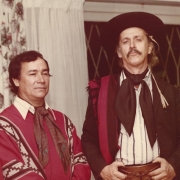 Ao fundo, uma janela com grade e cortinas branca. No centro da imagem há 4 quatro homens. No canto esquerdo, um homem branco, de cabelos médios, vestindo boina, um lenço no pescoço e uma camiseta branca. Ao seu lado, um homem branco, de cabelos curtos, vestindo um lenço no pescoço e um moletom vermelho com listras brancas. Em seu lado direito, Jayme Caetano Braun, um homem branco, de cabelos médios, vestindo roupas características da cultura gaúcha, como chapéu, lenço e bombacha. No canto direito, ao lado de Jayme, um homem branco, de cabelos curtos, vestindo um moletom preto.