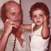 Na imagen, no lado esquerdo, Jayme Caetano Braun, um homem branco, de cabelos curtos, vestindo uma camiseta verde, segurando um telefone. Em seu colo, seu neto, Luciano, um menino de cabelos curtos, vestindo uma camiseta branca.