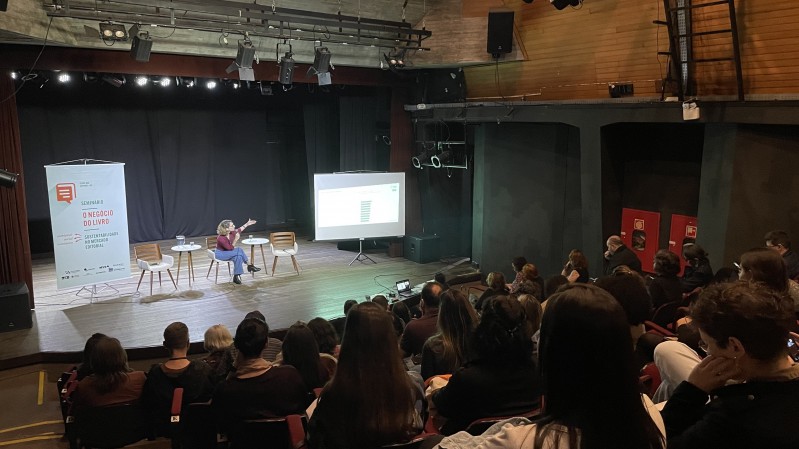 No palco de um teatro uma mulher sentada fala ao microfone enquanto aponta para uma tela, e a plateia assiste