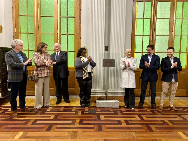 Sete pessoas batem palmas ao lado da placa de homenagem a Gilberto Schwartsmann 