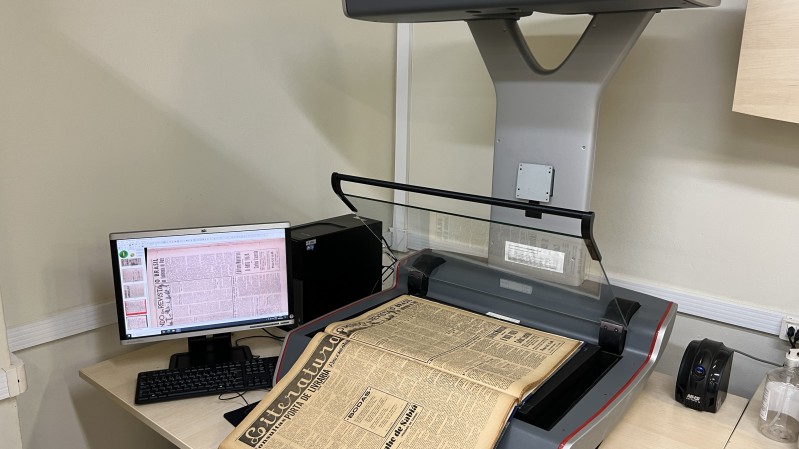 Sobre uma mesa, um jornal antigo é escaneado, enquanto a imagem escaneada aparece em um monitor de computador