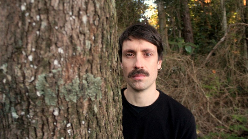 Foto de Pedro Longes. Ele é mostrado do peito para cima. É um homem branco, de cabelo e bigodes pretos, veste blusa preta e está olhando para a câmera, em pé, ao lado de uma árvore e em meio a outras árvores