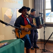 Um homem canta e toca violão e outro, ao seu lado, toca gaita. Ambos estão vestidos com trajes gaúchos