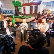 Beztriz Araujo rodeado de jornalistas, câmeras e microfones dando entrevista. Ao fundo, há uma alegoria do desfile já preparada para o desfile.
