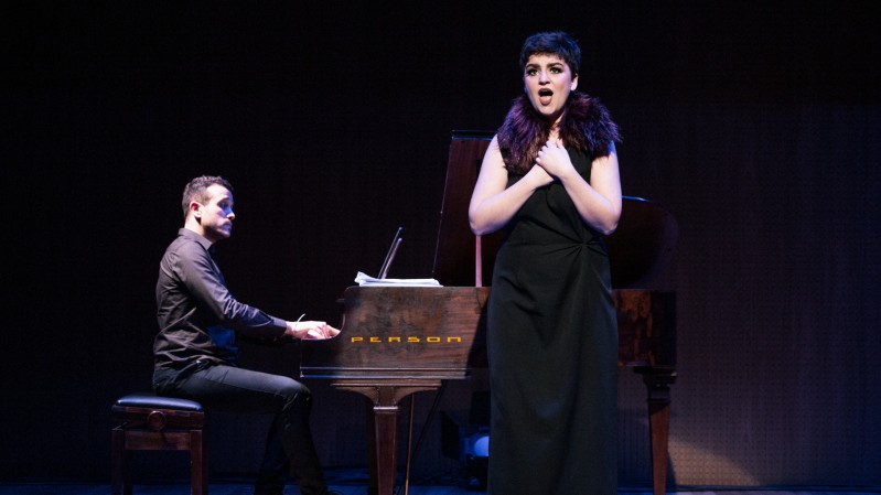 Na imagem, Jou Bennemann, uma mulher branca, de cabelos curtos, vestindo um vestido preto, cantando no 1º Recital Vozes do Ópera Estúdio.