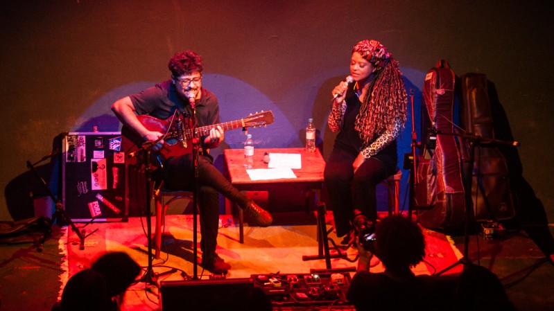 Na imagem, em um palco, Maurilio, um homem branco, de cabelos curtos, usando óculos, vestindo uma camisa e calça preta, tocando um violão sentado e cantando. Em seu lado, à direita, Nina Fola, uma mulher negra, de tranças longas, vestindo uma camisa e calça preta, cantando sentada.