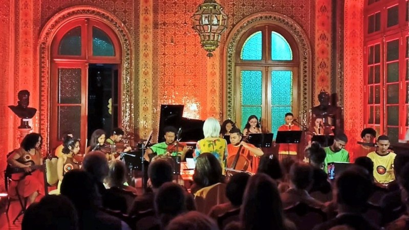 Imagem da Orquestra Villa-Lobos, na Biblioteca Pública do Estado (BPE), um concerto com jovens da Lomba do Pinheiro tocando instrumentos com cordas, com uma plateia assistindo ao show.