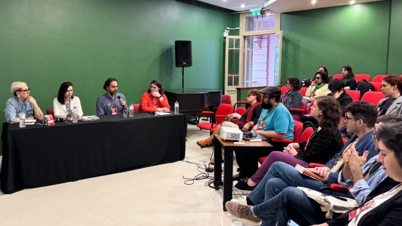 Imagem da mesa-redonda "Literatura e o Antropoceno", ambientada em uma sala com paredes verdes, composta por Natalia Borges Polesso, uma mulher branca, de cabelos curtos, usando óculos e camisa jenas azul, em sua direita, Morgana Kretzzman, uma mulher branca, de cabelos médios, vestindo um blazer branco, ao seu lado direito, Daniel Galera, um homem branco, de cabelos curtos, com barba grisalha, vestindo uma camiseta azul-marinho, em seu lado direito, na ponta da mesa, Andrezza Postay, uma mulher branca, de cabelos médios, vestindo óculos e blusa laranja. À frente da mesa, uma plateia assiste ao debate.