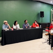 Imagem da mesa-redonda "Literatura e o Antropoceno", ambientada em uma sala com paredes verdes, composta por Natalia Borges Polesso, uma mulher branca, de cabelos curtos, usando óculos e camisa jenas azul, em sua direita, Morgana Kretzzman, uma mulher branca, de cabelos médios, vestindo um blazer branco, ao seu lado direito, Daniel Galera, um homem branco, de cabelos curtos, com barba grisalha, vestindo uma camiseta azul-marinho, em seu lado direito, na ponta da mesa, Andrezza Postay, uma mulher branca, de cabelos médios, vestindo óculos e blusa laranja. À frente da mesa, uma plateia assiste ao debate.