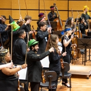 A imagem mostra uma orquestra de músicos em pé, ocupando o palco de uma sala de concerto. Todos os músicos estão vestidos de preto, mas muitos usam adereços coloridos e divertidos, como chapéus, tiaras, laços e faixas na cabeça. No canto esquerdo, alguns músicos usam chapéus de festa e acessórios floridos. No centro, há músicos com gravatas e chapéus verdes, além de uma mulher com uma saia preta e adereços floridos na cabeça. À direita, há músicos de cabelo colorido e faixas vermelhas. Todos seguram seus arcos levantados, como se tivessem terminado de tocar, em sinal de conclusão ou celebração.