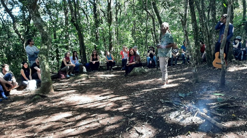 Na imagem, um grupo de pessoas está reunido em um ambiente de mata, sentadas no chão entre raízes e folhas caídas. Elas estão espalhadas entre as árvores altas e troncos finos, observando com interesse algo à sua frente. No centro da cena, um homem de pele negra e cabelos compridos presos está de pé, vestindo uma blusa estampada verde e calça clara, falando com o grupo. Perto dele, há uma fogueira apagada, com galhos dispostos em formato de estrela. À direita, outra pessoa está em pé, encostada em uma árvore, segurando a testa com a mão, enquanto uma guitarra está apoiada no tronco ao lado. Ao fundo, mais pessoas sentadas assistem atentamente à cena.