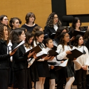 No canto superior esquerdo, várias jovens cantoras estão de pé, segurando pastas pretas abertas com partituras. No centro, há uma mistura de crianças e adolescentes, todas vestindo roupas formais, predominantemente brancas com detalhes pretos, como gravatas e laços. No canto direito, mais jovens continuam o grupo, algumas delas usando óculos, todas igualmente segurando suas partituras enquanto cantam. Ao fundo, as paredes têm um tom amadeirado claro. 