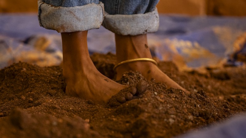 Na parte superior da imagem, é possível ver um fundo levemente desfocado com tons de marrom e azul, sugerindo um ambiente ao ar livre com chão de terra. Centralizado, há uma pessoa negra com calças jeans dobradas na altura dos tornozelos, destacando seus pés descalços tocando o solo. O pé direito está parcialmente coberto por terra, enquanto o esquerdo está mais à mostra. Ao redor do tornozelo esquerdo, há uma tornozeleira dourada. A terra é de cor marrom avermelhada.