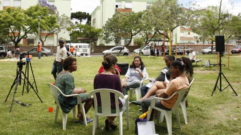 A imagem é ambientada em uma praça, com brinquedos e gramado verde. No centro da imagem está um grupo de sete mulheres, sentadas em cadeiras brancas, com microfone e uma câmera.