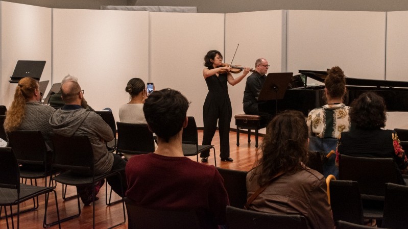 Em um salão com paredes de cor clara e painéis brancos ao fundo, uma violinista e um pianista se apresentam no centro da imagem. A violinista está em pé, vestida com um macacão preto, enquanto toca o violino. Ao lado, sentado em um banco, o pianista está vestido de preto, tocando piano. A plateia, em cadeiras dispostas em fileiras, observa a apresentação. Algumas pessoas assistem atentamente. O chão do salão é de madeira.