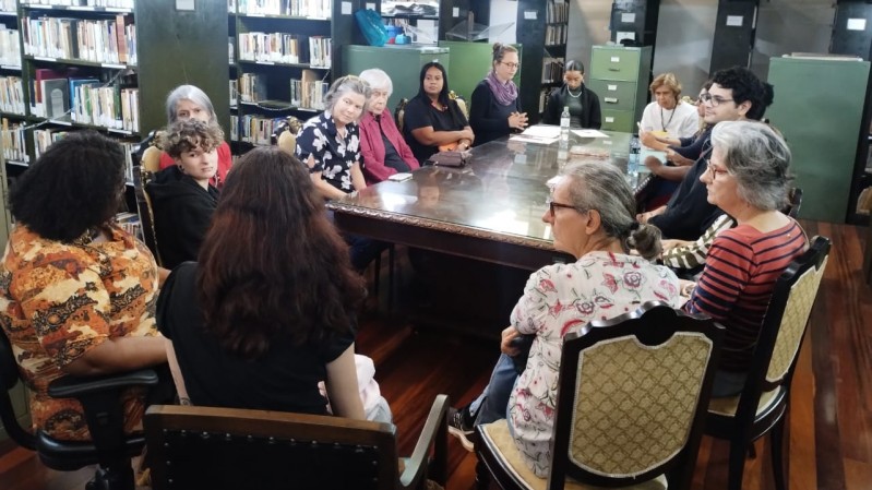 A imagem mostra uma reunião de um grupo de pessoas na Biblioteca Pública do Estado (BPE). Ao fundo, há prateleiras altas repletas de livros organizados. Acima das estantes, há uma estrutura metálica com iluminação, e no centro da sala, há uma mesa retangular grande e de madeira escura, onde as pessoas estão sentadas ao redor. Na mesa, há cerca de 14 pessoas, de diferentes idades, sentadas em cadeiras de estilo antigo. O piso é de madeira.