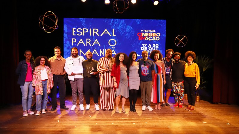 Treze pessoas em pé, sobre o palco. São premiados, curadores e equipe do Festival