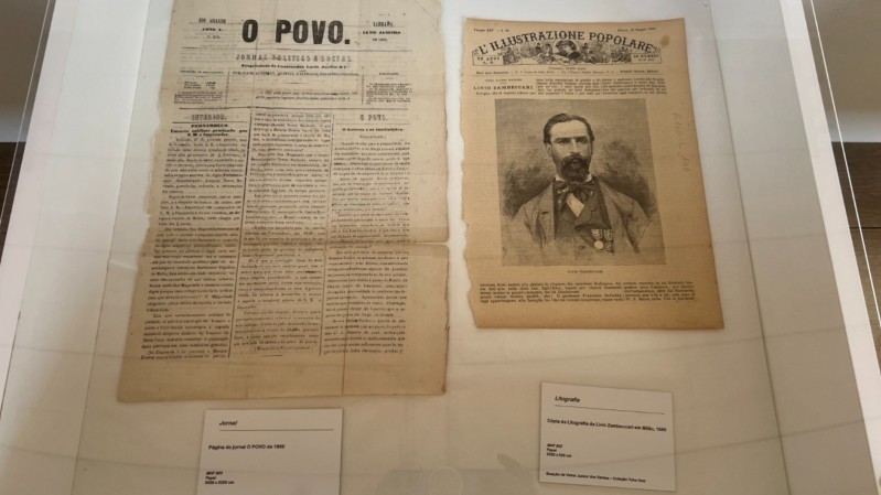 A imagem mostra duas páginas de publicações antigas, em um fundo branco, dispostas lado a lado e protegidas por um vidro transparente. À esquerda, está uma edição do jornal "O Povo", datada de 1860, com layout em colunas, texto denso em preto sobre papel envelhecido. À direita, há uma página de uma publicação italiana, com o título "L'Illustrazione Popolare" no topo, acompanhada por uma imagem central de Livio Zambeccari, um homem com barba e cabelos curtos, vestido com terno e medalhas. Abaixo desta página, uma outra placa de identificação descreve: "Litografia – Cópia da Litografia de Livio Zambeccari em Milão, 1868".