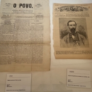 A imagem mostra duas páginas de publicações antigas, em um fundo branco, dispostas lado a lado e protegidas por um vidro transparente. À esquerda, está uma edição do jornal "O Povo", datada de 1860, com layout em colunas, texto denso em preto sobre papel envelhecido. À direita, há uma página de uma publicação italiana, com o título "L'Illustrazione Popolare" no topo, acompanhada por uma imagem central de Livio Zambeccari, um homem com barba e cabelos curtos, vestido com terno e medalhas. Abaixo desta página, uma outra placa de identificação descreve: "Litografia – Cópia da Litografia de Livio Zambeccari em Milão, 1868".