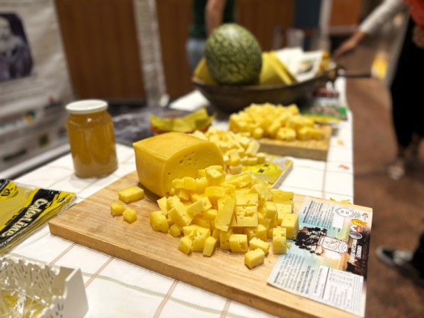 Imagem mostra uma mesa com toalha de estampa xadrez, onde está servida uma tábua com Queijo Artesanal Serrano em cubos pequenos e uma peça maior. Ao longo da mesa, se vê outras duas tábuas com o queijo e, em desfoque, uma cexta com diversos queijos.