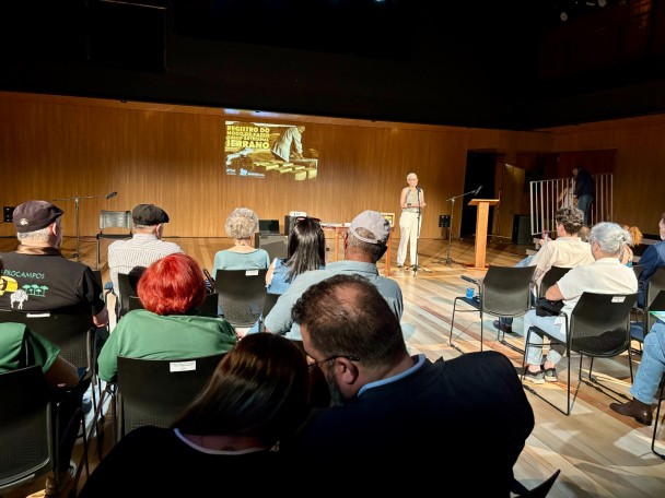 A imagem mostra o Teatro Olga Reverbel, todo revestido em madeira, em plano aberto. Projetado na parede mais distante da imagem, uma imagem de uma pessoa de costas manuseando peças inteiras de queijos e o título alinhado à esquerda: “Registro do Modo de Fazer do Queijo Artesanal Serrano”, logo abaixo estão os logos do IPHAE e da Sedac. No palco, a Secretária de Estado da Cultura, Beatriz Araujo está ao microfone diante de diversas pessoas posicionadas em platéia. 