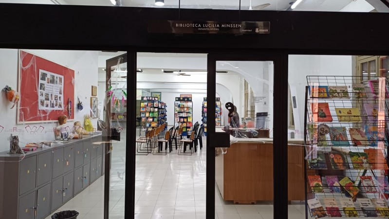 Imagem de entrada da Biblioteca Lucília Minssen. No centro, uma porta de vidro onde é exposto o interior do espaço, com uma bancada de madeira marrom à direita, e ao fundo estandes de livros e cadeiras.