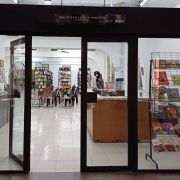 Imagem de entrada da Biblioteca Lucília Minssen. No centro, uma porta de vidro onde é exposto o interior do espaço, com uma bancada de madeira marrom à direita, e ao fundo estandes de livros e cadeiras.