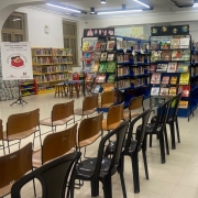 Imagem do interior da Biblioteca Lucília Minssen, com três estandes azuis com livros, um banner com o logo da instituição e cadeiras marrons e pretas vazias.