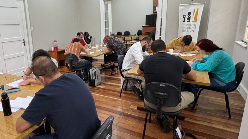 Em uma sala de tom claro de cinza e com piso de madeira, diversas pessoas estão dispostas em três mesas de reunião escrevendo a mão.