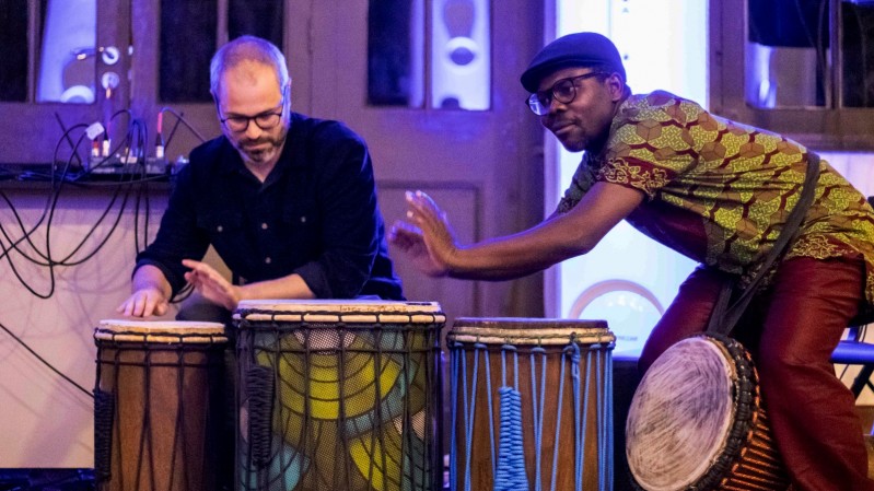 No fundo, há uma parede com portas de vidro emolduradas por madeira, que refletem luzes azuladas e objetos difusos. No centro da imagem, dois homens estão tocando tambores africanos. À esquerda, Felipe Merker Castellani, um homem de óculos, pele clara e barba grisalha, usando uma camisa preta e tocando um tambor com as mãos. À direita, Loua Pacom Ouali, um homem de pele negra, óculos e boné preto, vestindo uma camisa estampada com padrões geométricos verde e vermelho, tocando um tambor com a mão direita. 