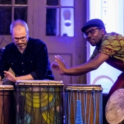 No fundo, há uma parede com portas de vidro emolduradas por madeira, que refletem luzes azuladas e objetos difusos. No centro da imagem, dois homens estão tocando tambores africanos. À esquerda, Felipe Merker Castellani, um homem de óculos, pele clara e barba grisalha, usando uma camisa preta e tocando um tambor com as mãos. À direita, Loua Pacom Ouali, um homem de pele negra, óculos e boné preto, vestindo uma camisa estampada com padrões geométricos verde e vermelho, tocando um tambor com a mão direita. 